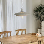 Sheer curtains and a tall palm frame a white Clérachic pendant hanging over a light wooden dining table.