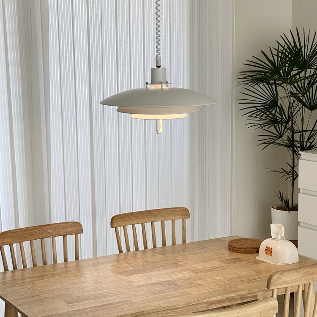 Sheer curtains and a tall palm frame a white Clérachic pendant hanging over a light wooden dining table.