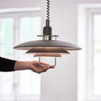 A hand holds the lower handle of this black Clérachic pendant, showing how the three tiers sit below a coiled cable.
