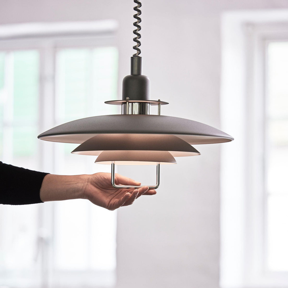 A hand holds the lower handle of this black Clérachic pendant, showing how the three tiers sit below a coiled cable.