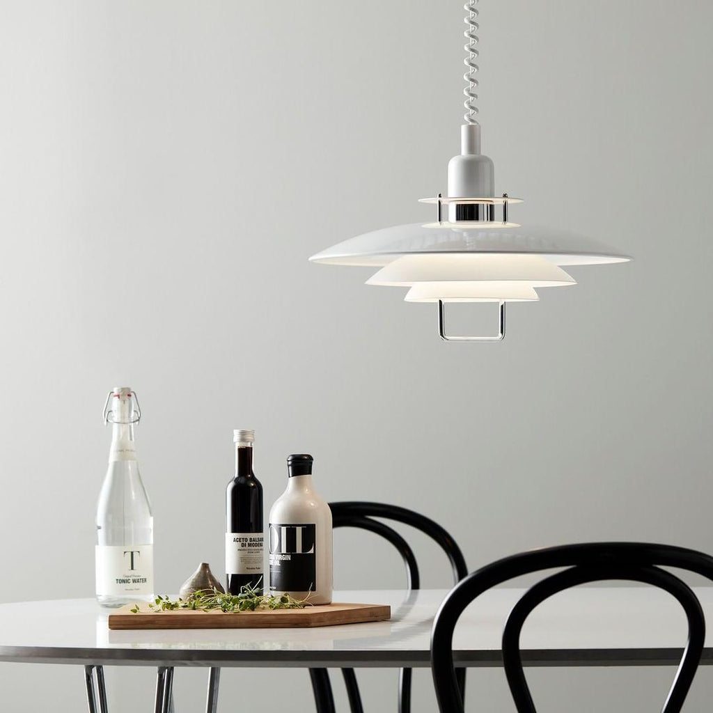 Over a white table holding oil, vinegar and a board of herbs, a white Clérachic pendant hangs near black bentwood chairs.