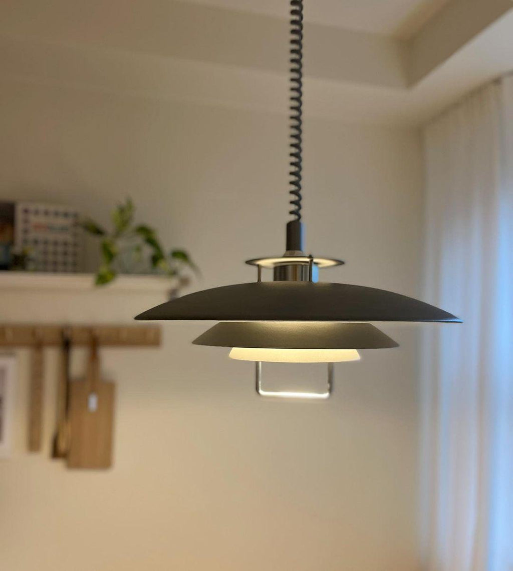 A black Clérachic pendant glows warmly in a kitchen, with a shelf of utensils and a trailing plant on the wall.