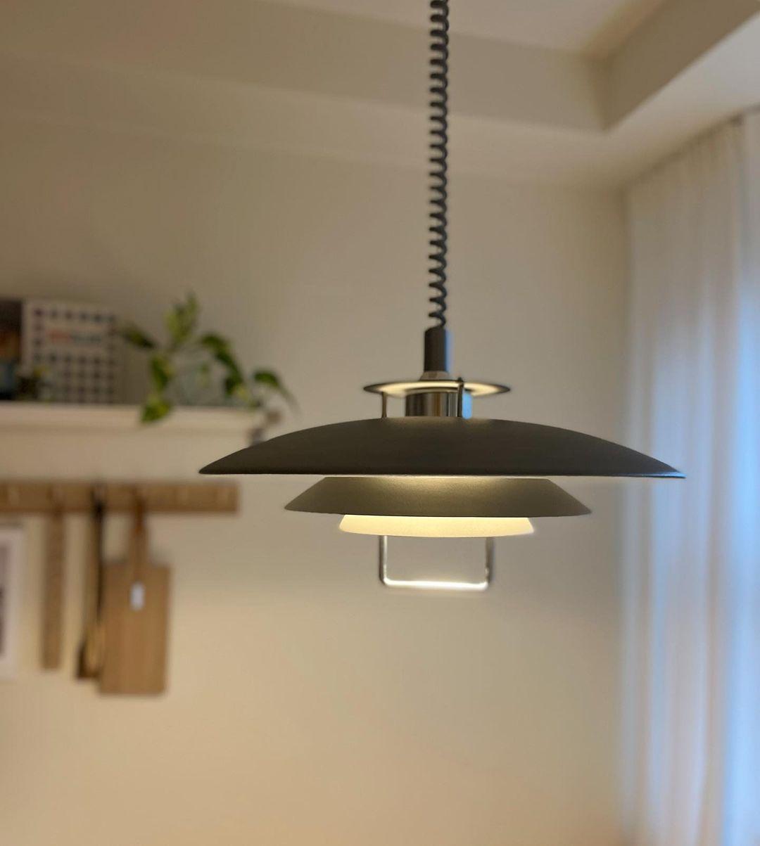 A black Clérachic pendant glows warmly in a kitchen, with a shelf of utensils and a trailing plant on the wall.