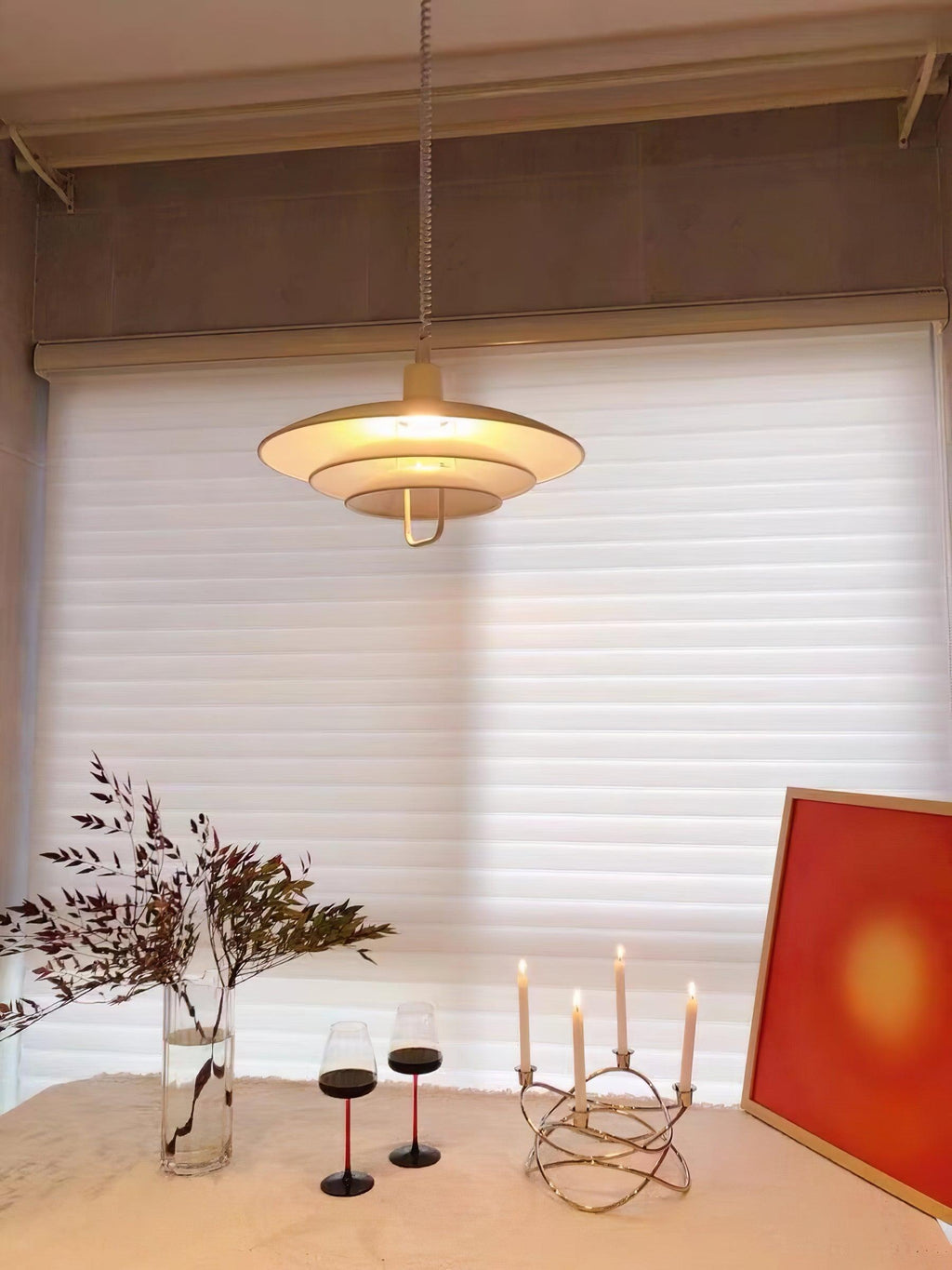 Candlelight and a red print sit below a warm-glowing Clérachic pendant, hung in front of white roller blinds.