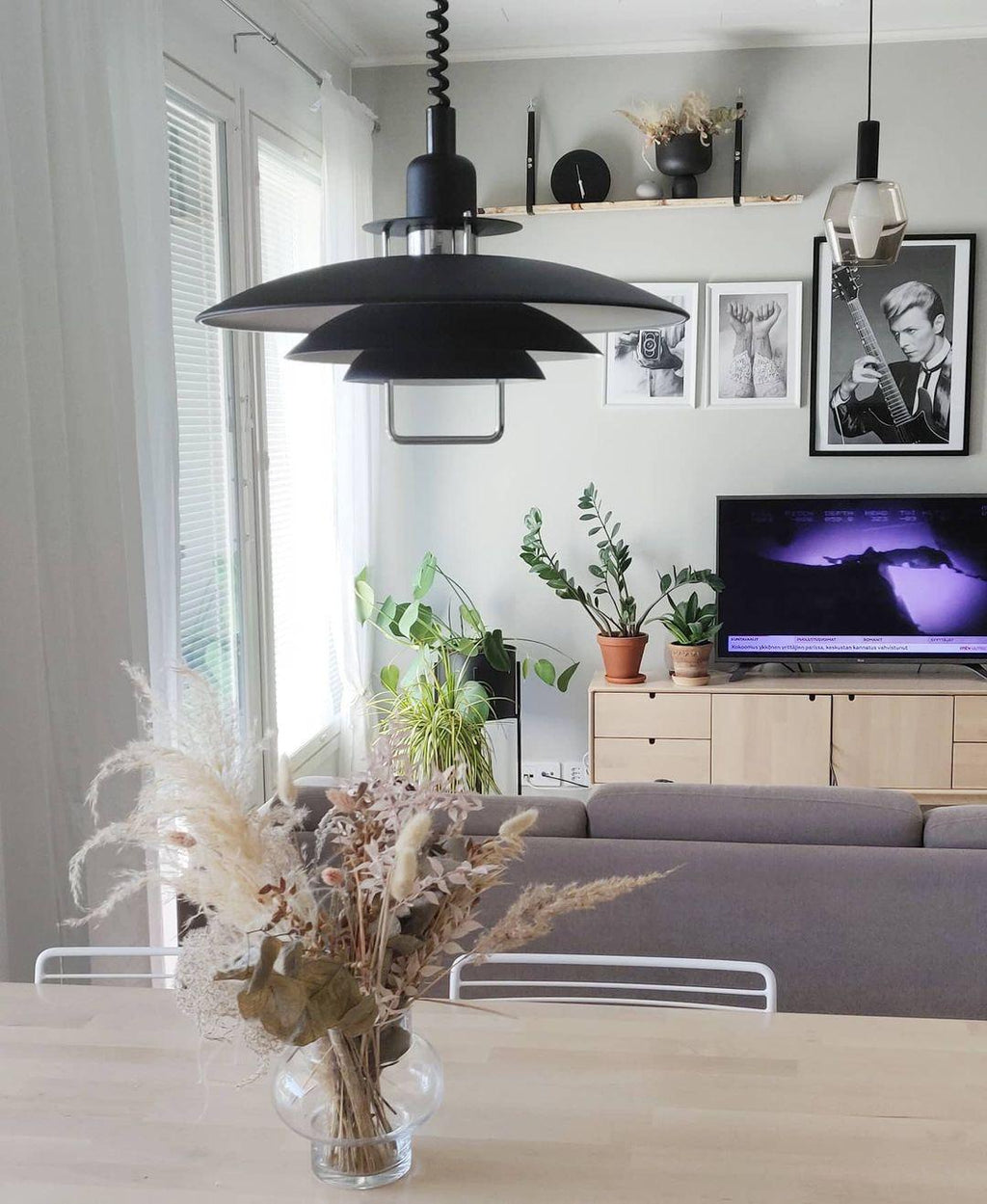 A black Clérachic pendant hangs over a pale wood table with dried grasses, in a living room with a sofa and a TV.