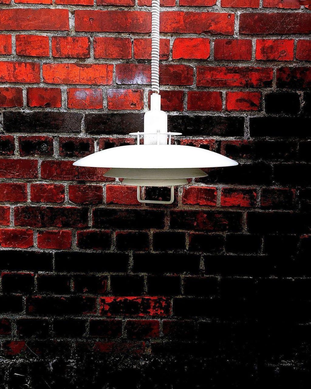 Against a dark red brick wall, a white Clérachic pendant stands out, its coiled cable dropping from above.