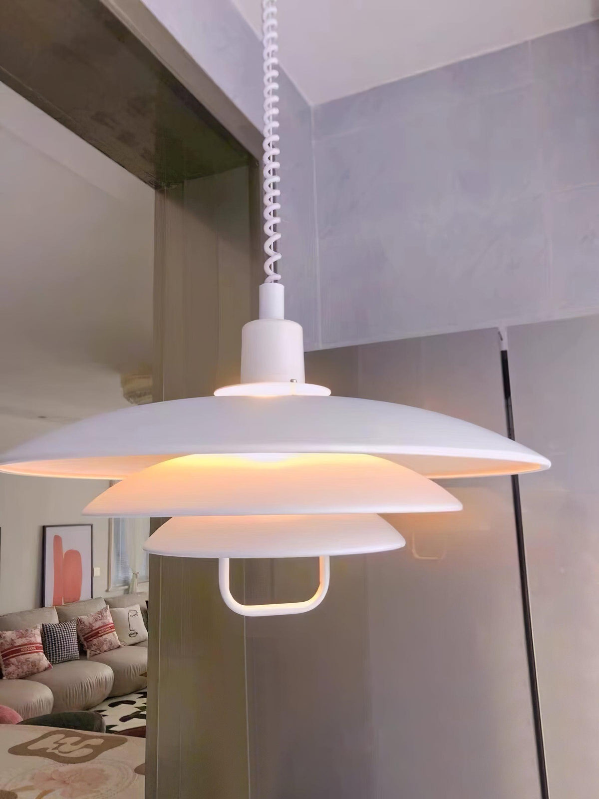 Warm light glows between the white tiers of this Clérachic pendant, seen from below with a living room blurred behind.