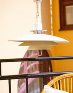 A white Clérachic pendant catches yellow light against an orange wall, with a black railing and a wicker chair below.