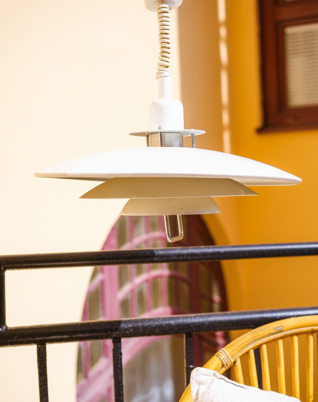 A white Clérachic pendant catches yellow light against an orange wall, with a black railing and a wicker chair below.