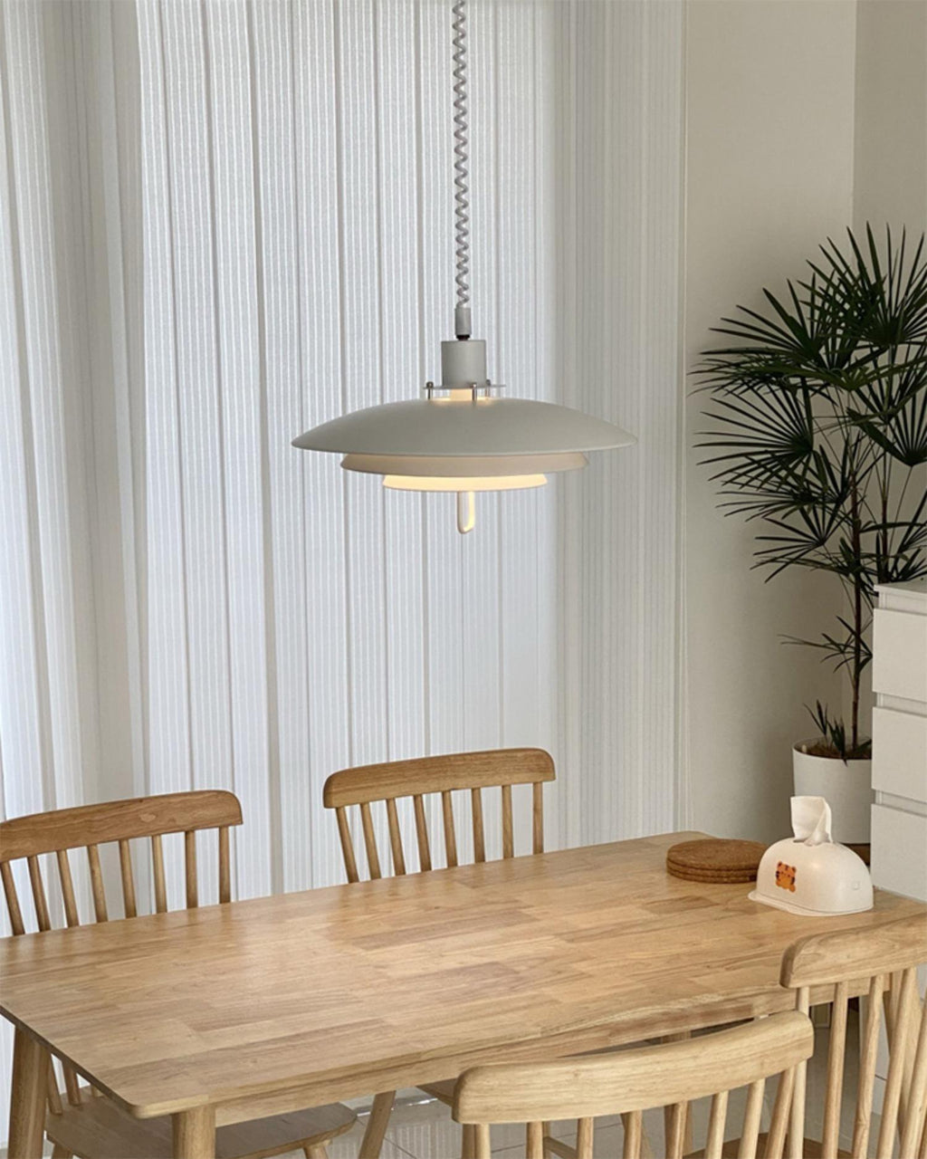 A white Clérachic pendant hangs on a coiled cable above a pine dining table with slatted chairs.