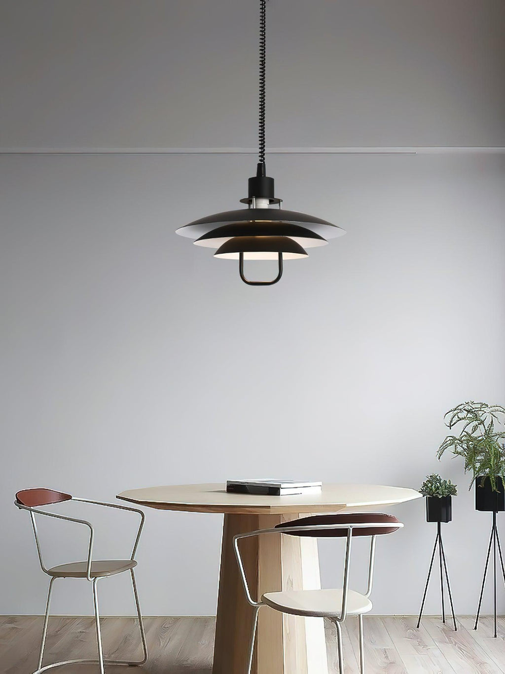 A black Clérachic pendant hangs above a round wooden table with two metal-framed chairs, against a plain grey wall.