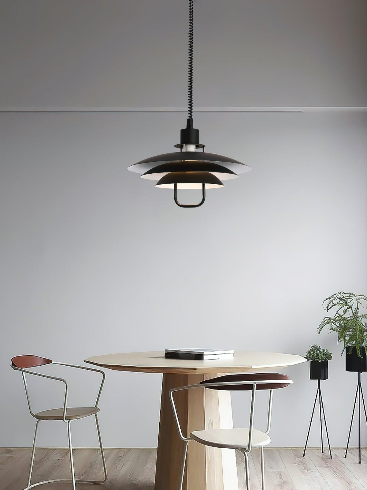 A black Clérachic pendant hangs above a round wooden table with two metal-framed chairs, against a plain grey wall.