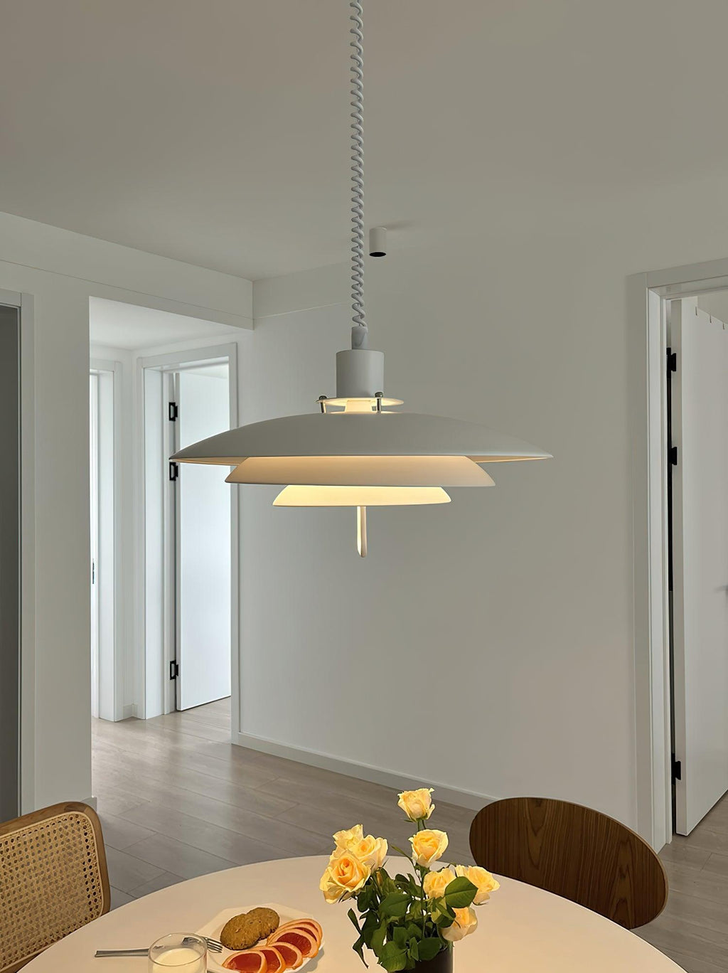 White Clérachic pendant glowing softly in a light hallway, above a round table with fruit and a bunch of roses.