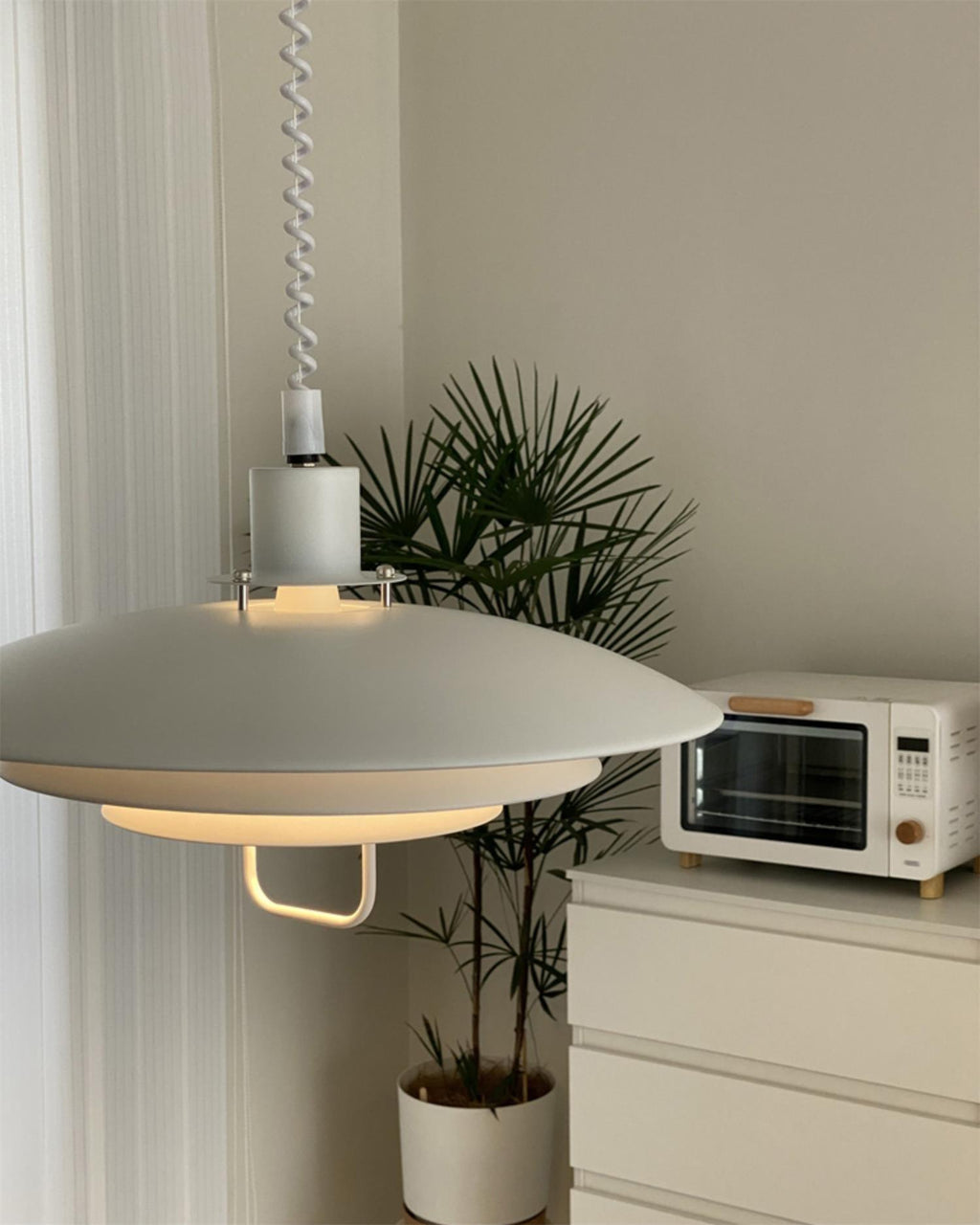 Close view of the broad white top shade on this Clérachic pendant, with a palm and a small oven on a chest of drawers beyond.