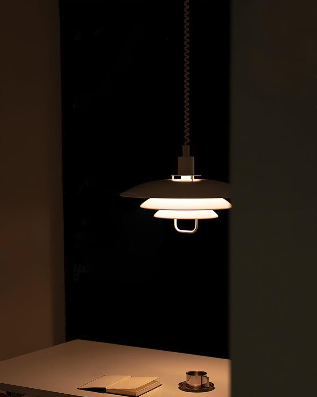 In a dark room, a Clérachic pendant lights a table with a notebook and a cup, its tiers glowing amber.