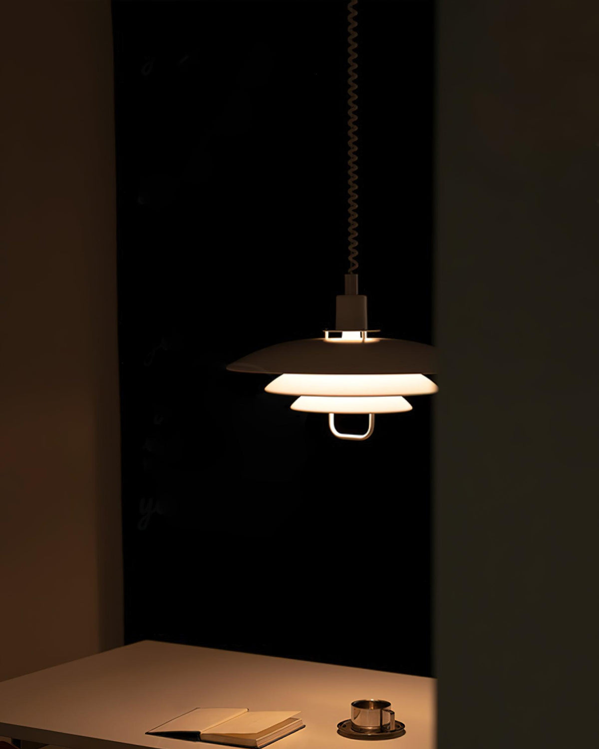 In a dark room, a Clérachic pendant lights a table with a notebook and a cup, its tiers glowing amber.