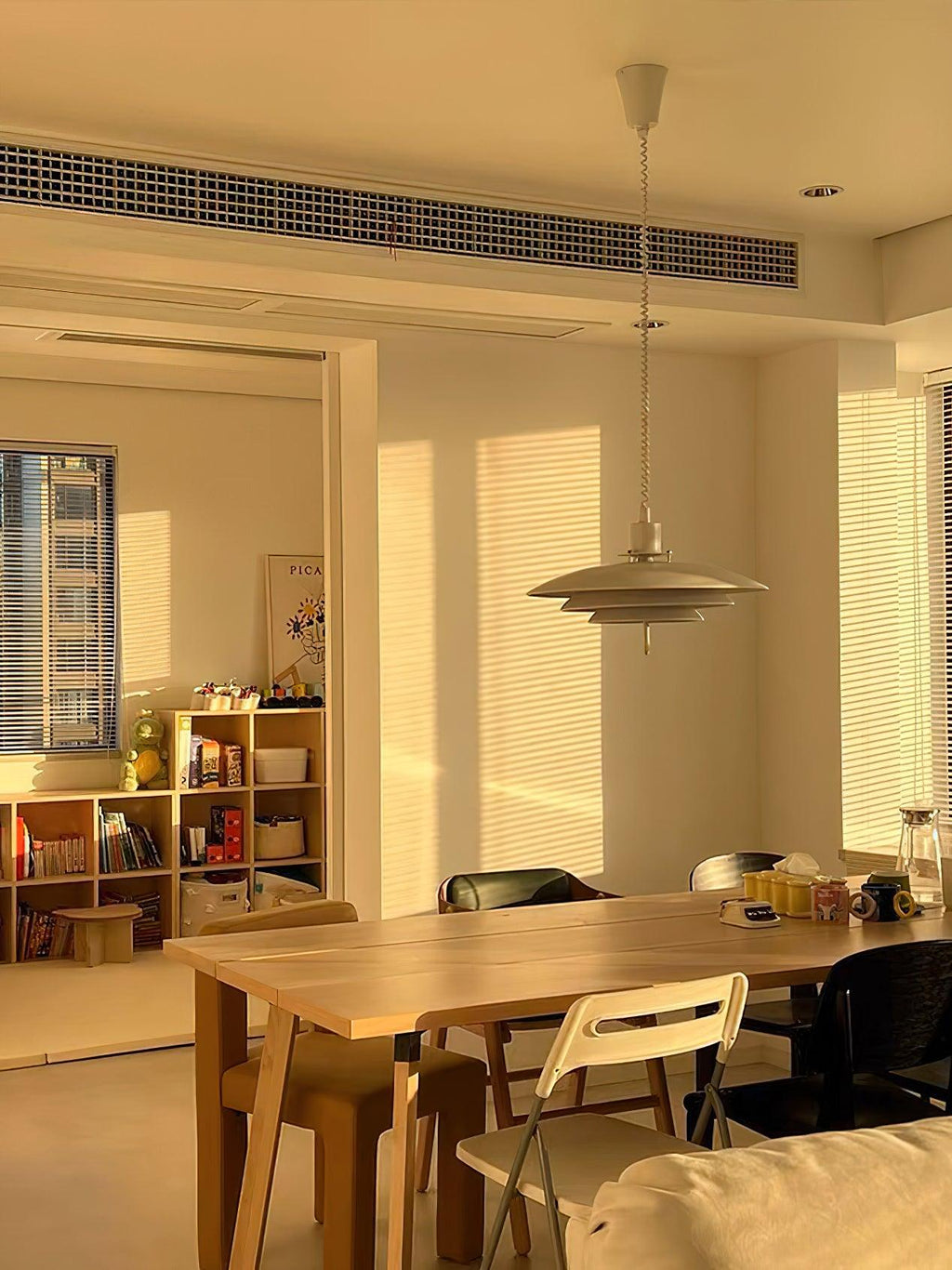 Late sun slants across a room where a Clérachic pendant hangs above a wooden table and a shelf of books.