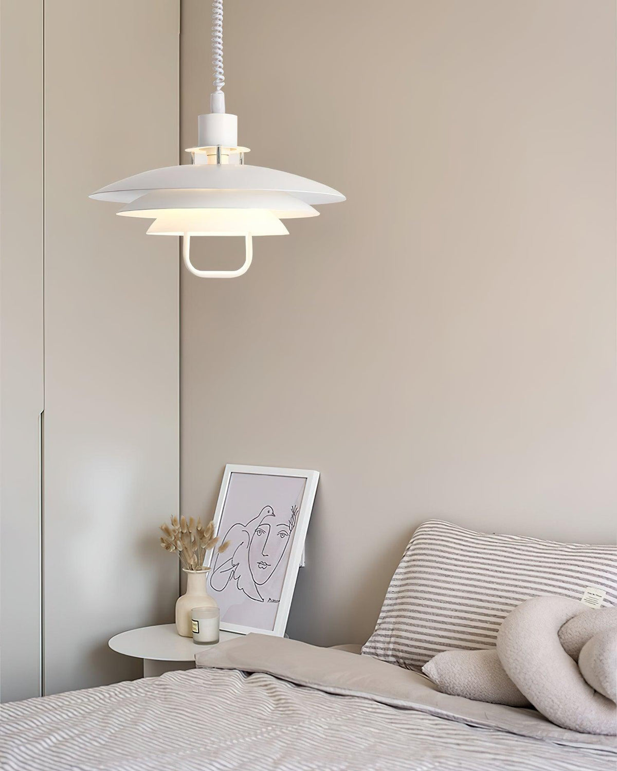 A white Clérachic pendant hangs beside a bed with striped bedding, a framed line drawing on the side table.
