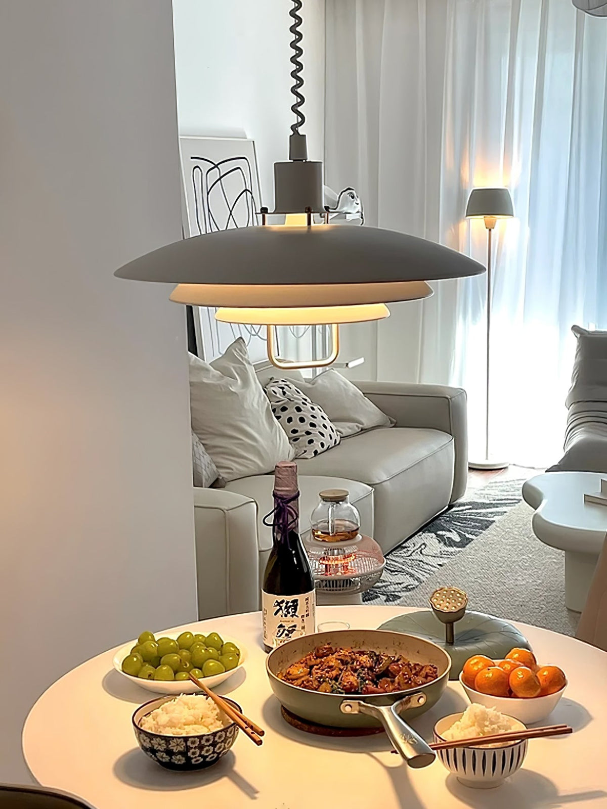 A pale grey Clérachic pendant glows above a round table laid with a meal, with a sofa and floor lamp behind.