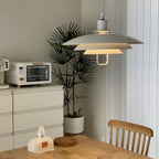 Tilted view of a white Clérachic pendant lit warm, above a pine table with a small oven and a palm in the corner.
