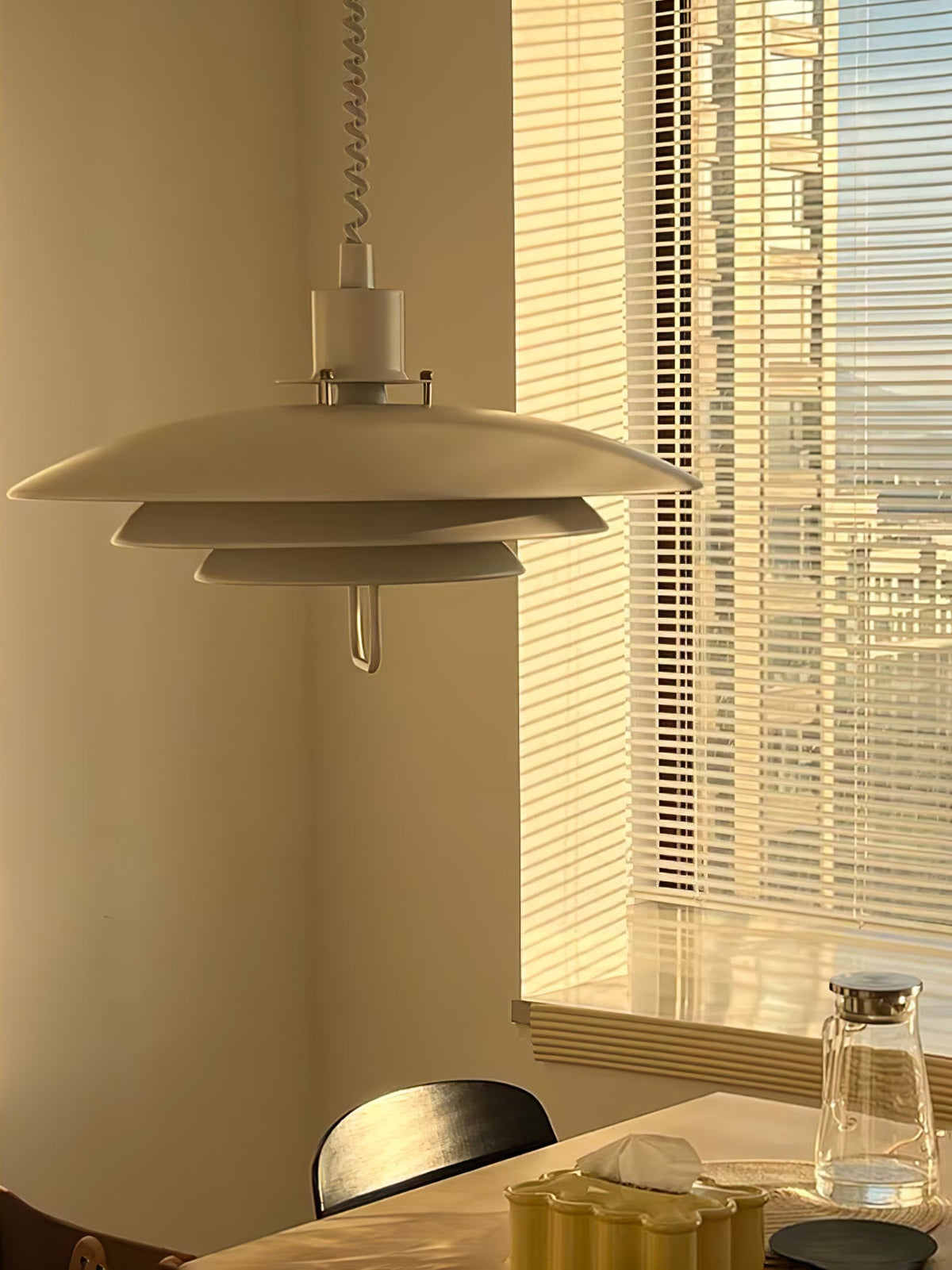 Golden light from window blinds falls on a pale Clérachic pendant, hung above a table with a glass jug.