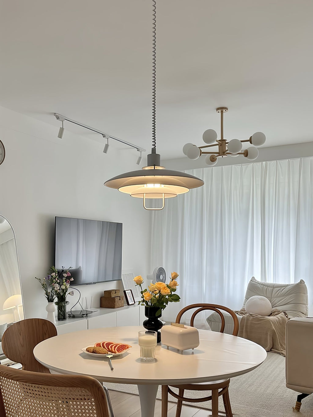 A Clérachic pendant hangs above a round white table in a living room with track lights and a white-curtained window.