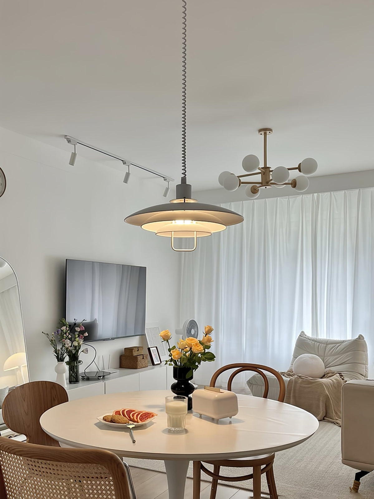 A Clérachic pendant hangs above a round white table in a living room with track lights and a white-curtained window.