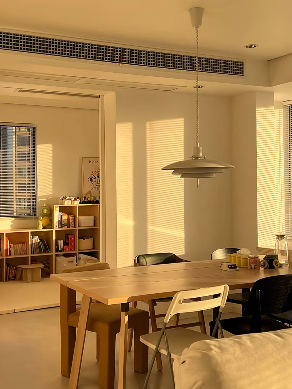 A Clérachic pendant hangs in a sunlit room, with striped light on the walls above a wooden table and folding chairs.