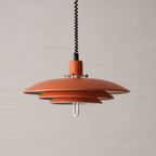 Orange Clérachic pendant with a black coiled cable and a white loop handle, hanging in front of a pale beige wall.