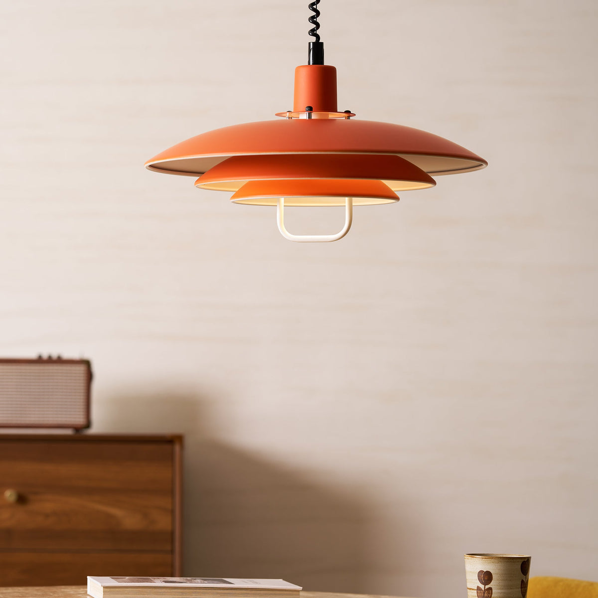Orange Clérachic pendant above a walnut cabinet with a small speaker, set against a pale wall.