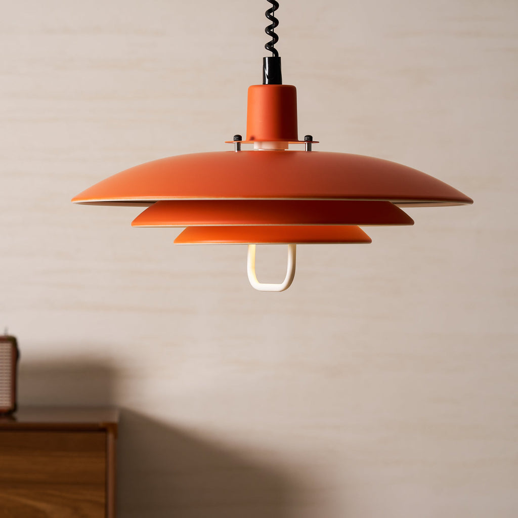 A wide orange Clérachic pendant hangs in front of a pale wall, with the black cable running up out of frame.