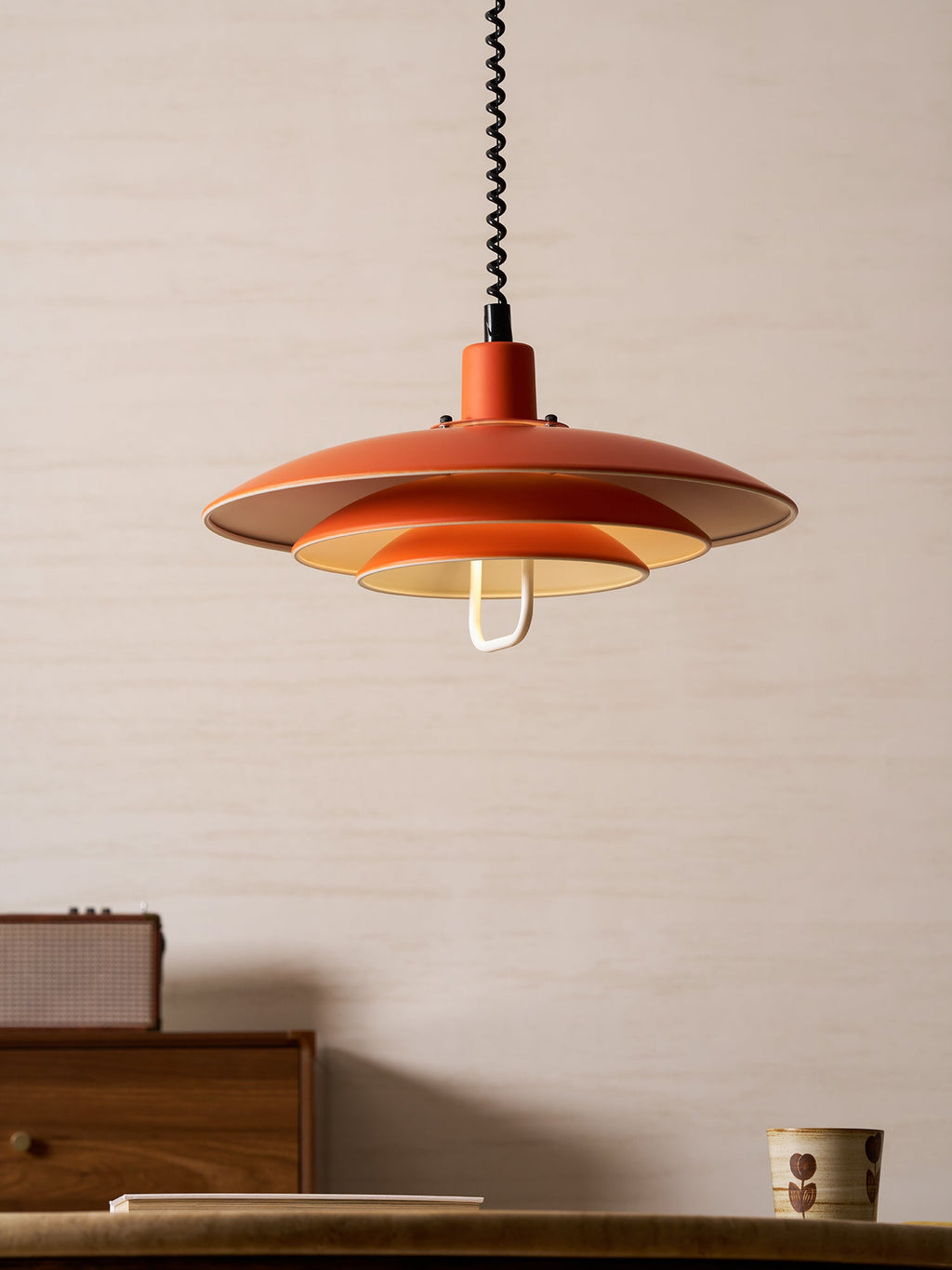 A neat orange Clérachic pendant hangs above a walnut drawer unit and a ceramic cup, with a pale wall behind.