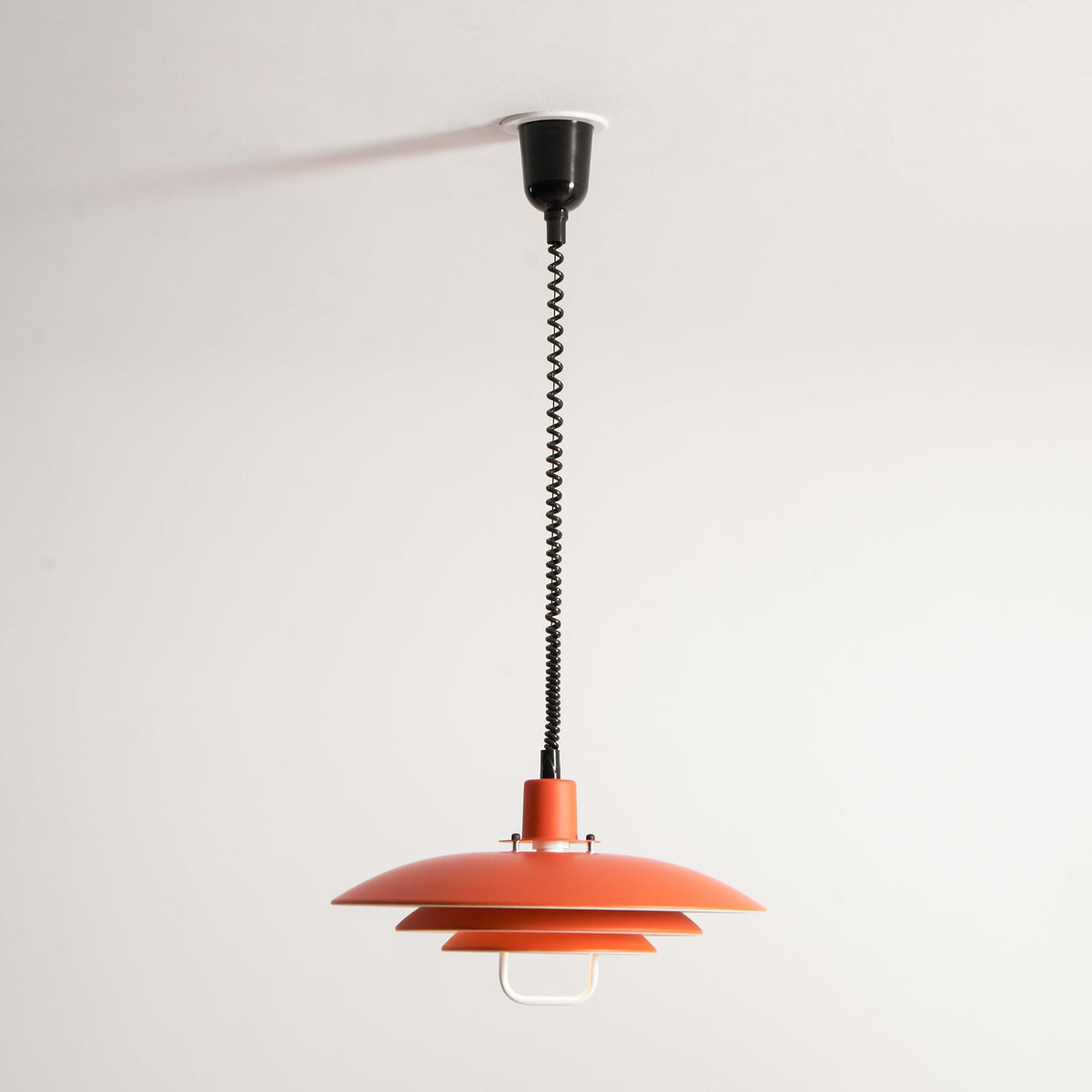 Orange Clérachic pendant hung on its full cable drop from a black ceiling cup, against a plain white wall.