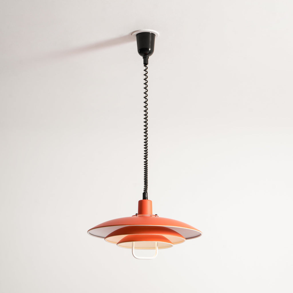 The same orange Clérachic pendant with its coiled cable shortened, from a black ceiling cup, lit inside.