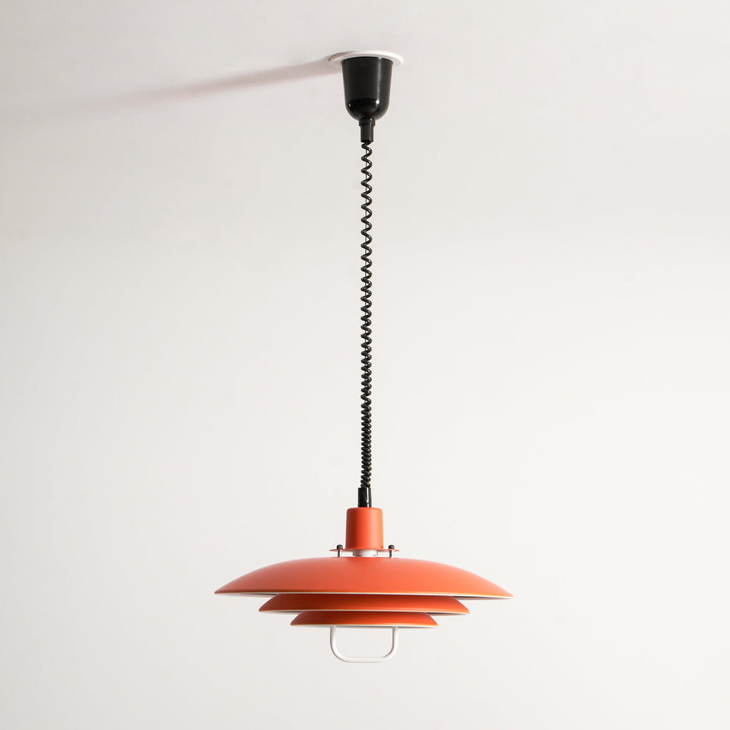 Orange Clérachic pendant in a high, long drop from a black ceiling cup, shown small against a white wall.