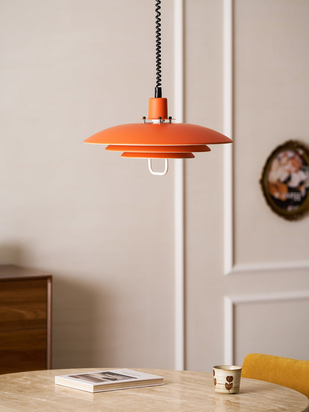 An orange Clérachic pendant hangs near a panelled door, above a stone-top table with a book and a cup.