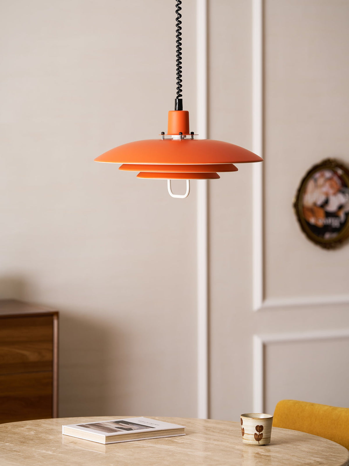 An orange Clérachic pendant hangs near a panelled door, above a stone-top table with a book and a cup.