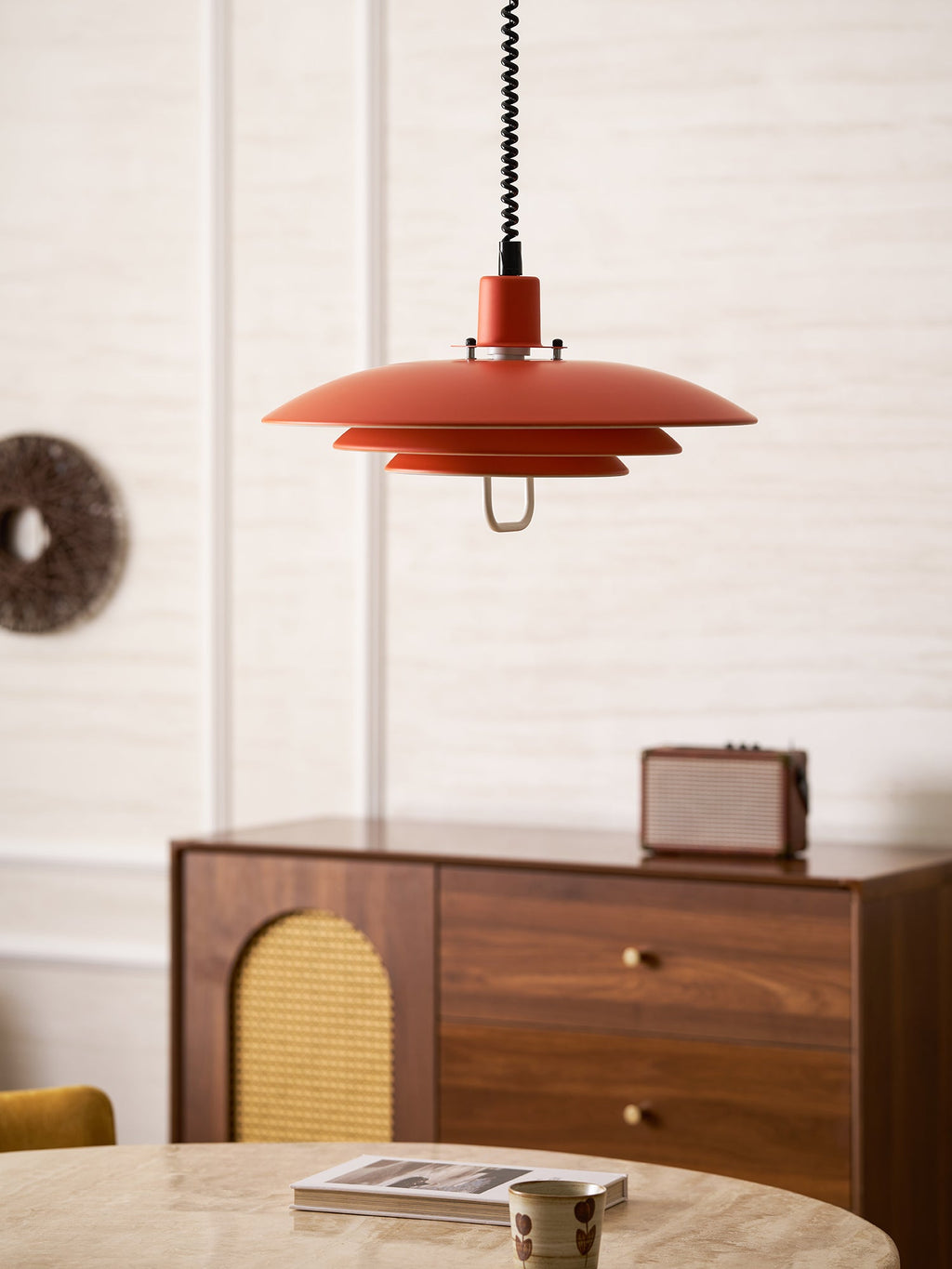 An orange Clérachic pendant hangs in front of a panelled wall, above a walnut sideboard with a rattan door.