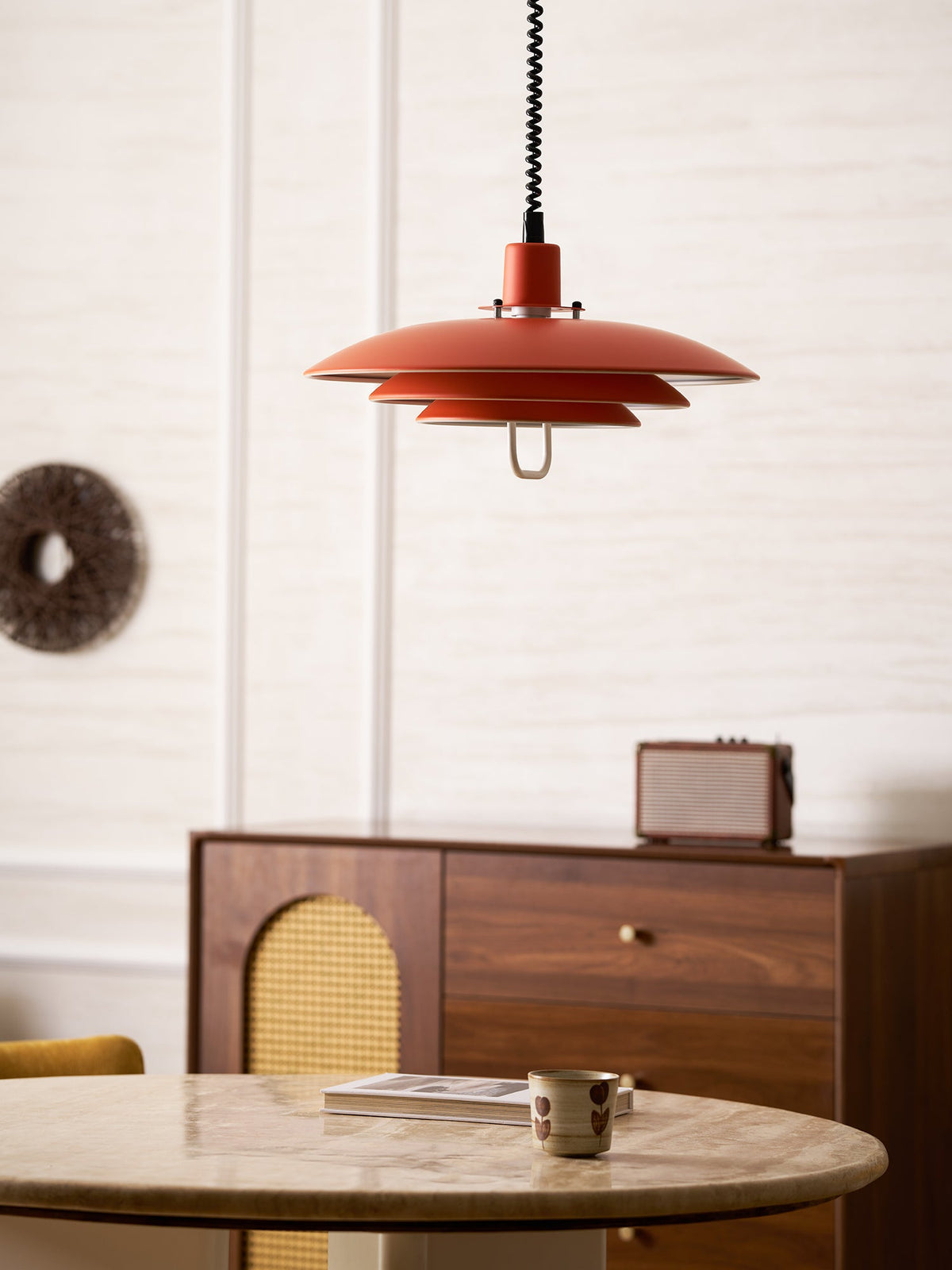 Orange Clérachic pendant seen from further back, with a round stone table, a rattan-fronted sideboard and a woven wall piece.