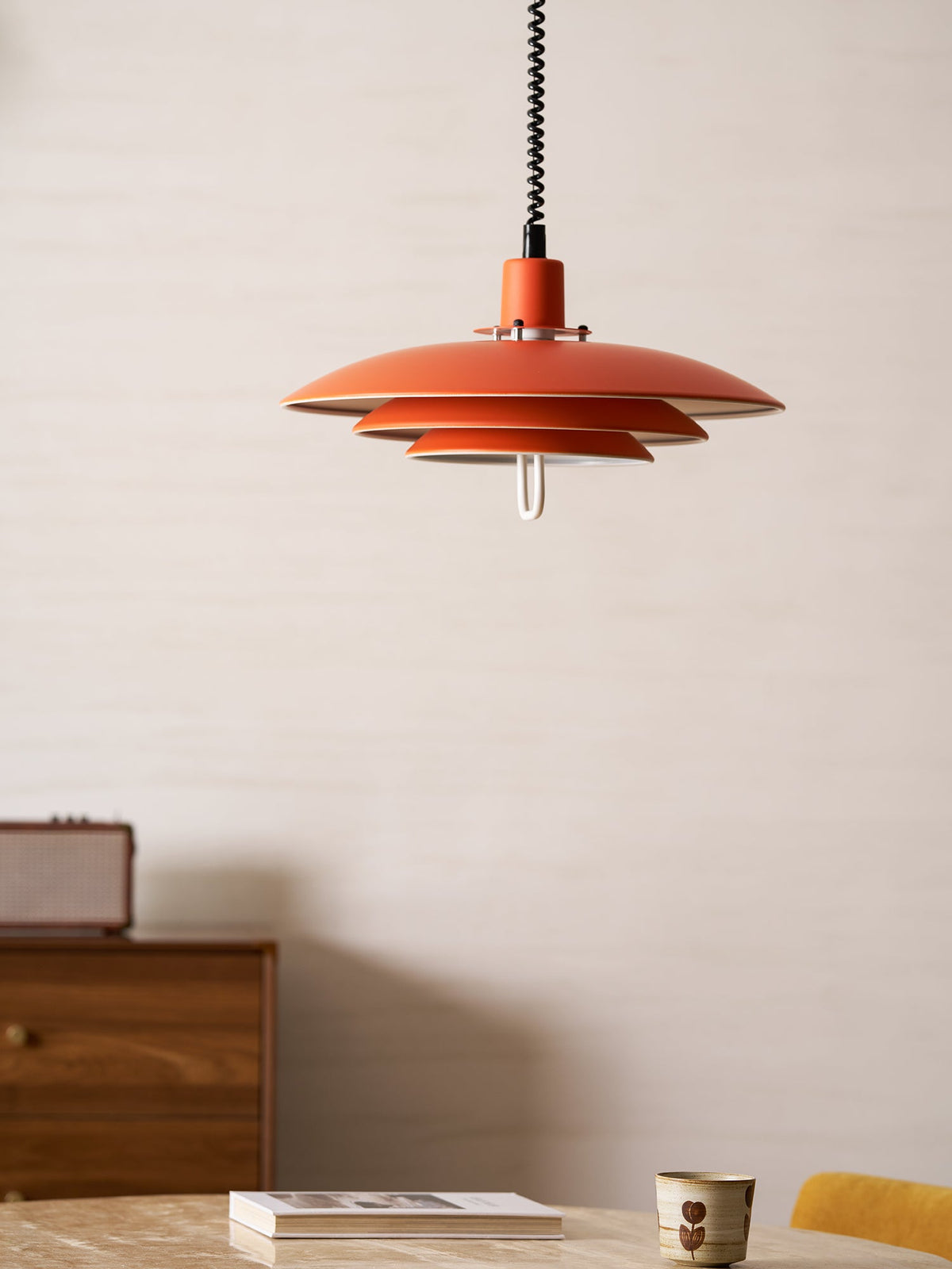 A close-framed orange Clérachic pendant, its three tiers hung over a sideboard with a table and a cup in view.