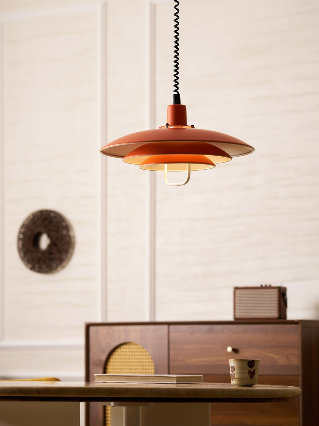 An orange Clérachic pendant hangs by a round woven wall piece, above a sideboard and a stone-top table.