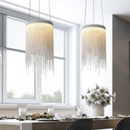 Two lit Clérachic chain-tassel pendants hang beside a tall window above a dining table set with flowers and glassware.
