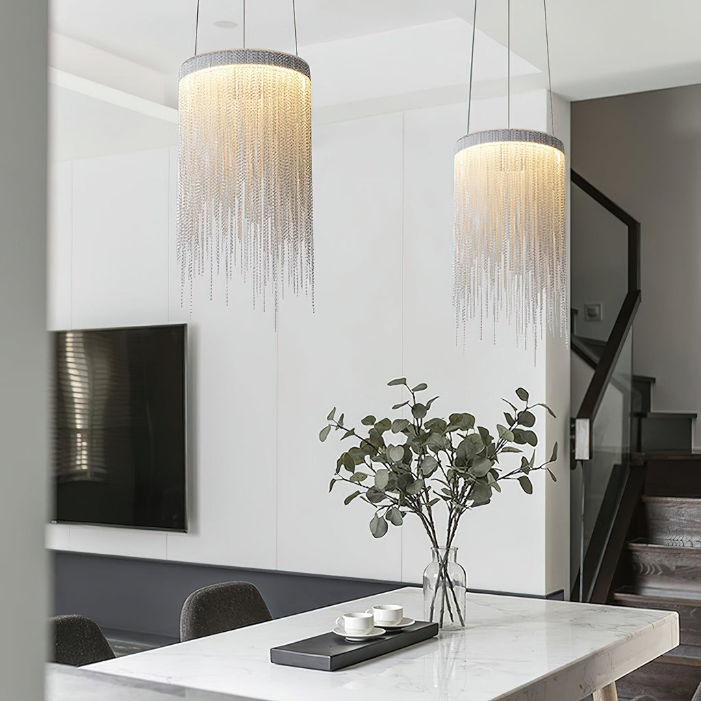A pair of Clérachic chain pendants at different heights in a bright living area, with a marble table and a plant below.