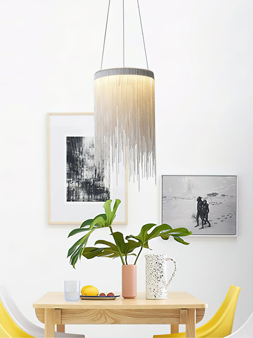 One Clérachic chain-tassel pendant over a small wooden table, with framed prints, a leafy plant and yellow chairs behind.