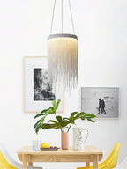 One Clérachic chain-tassel pendant over a small wooden table, with framed prints, a leafy plant and yellow chairs behind.