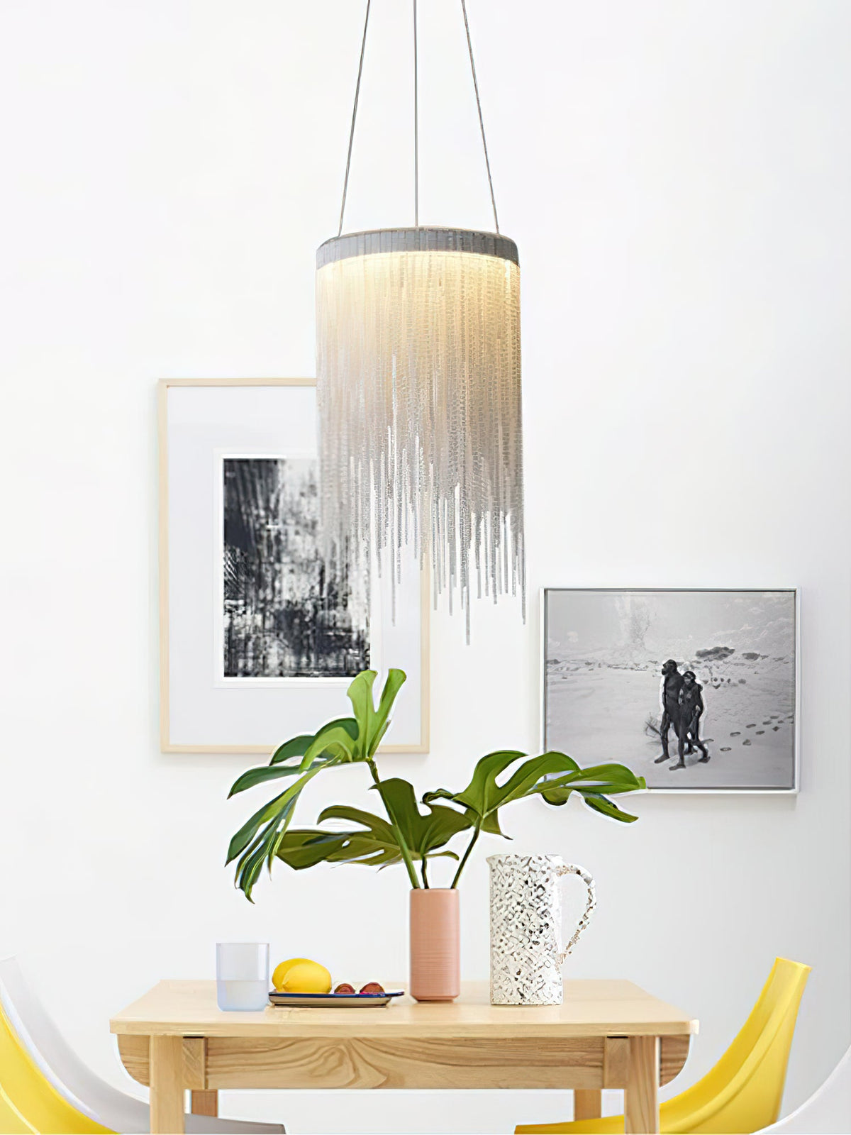 One Clérachic chain-tassel pendant over a small wooden table, with framed prints, a leafy plant and yellow chairs behind.