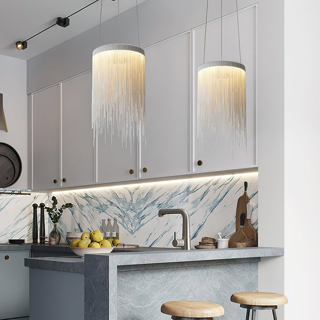 Two Clérachic chain pendants over a kitchen island with stools, in front of grey cabinets and a veined stone splashback.