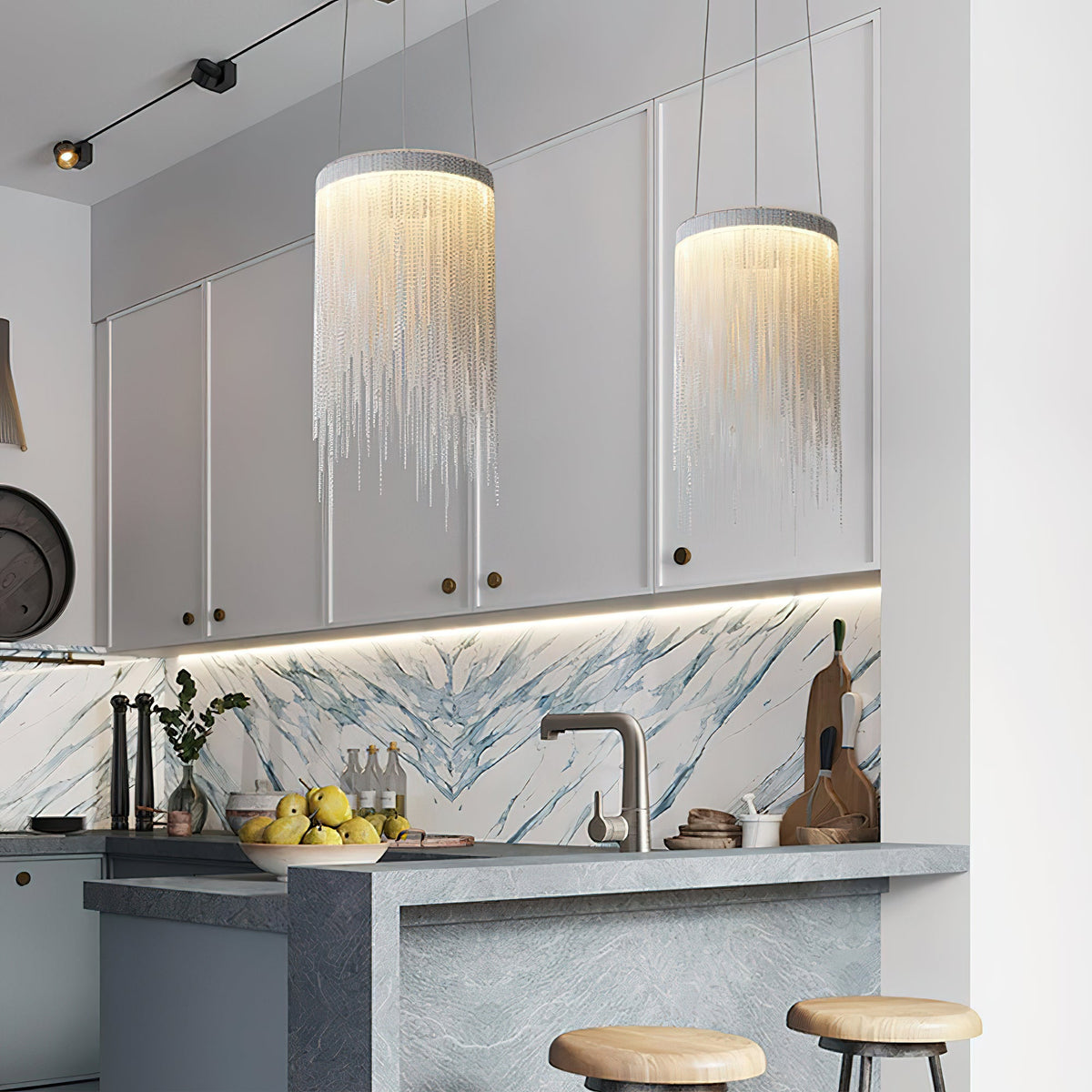 Two Clérachic chain pendants over a kitchen island with stools, in front of grey cabinets and a veined stone splashback.