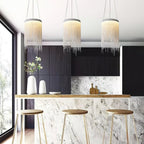 Three Clérachic chain pendants hang above a marble-fronted breakfast bar with wooden stools and a dark cabinet wall.