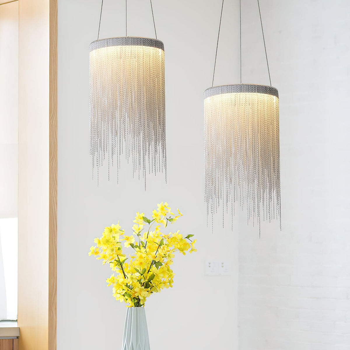 Two Clérachic chain-tassel pendants hang at staggered heights beside a vase of yellow flowers against a white brick wall.